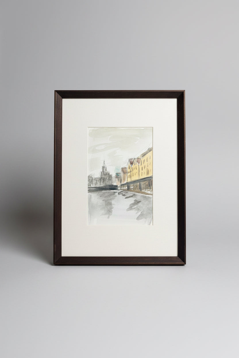 A framed watercolor painting of a city street scene with buildings and a church spire, matted with white and enclosed in a dark brown wooden frame, photographed on a gray tabletop.