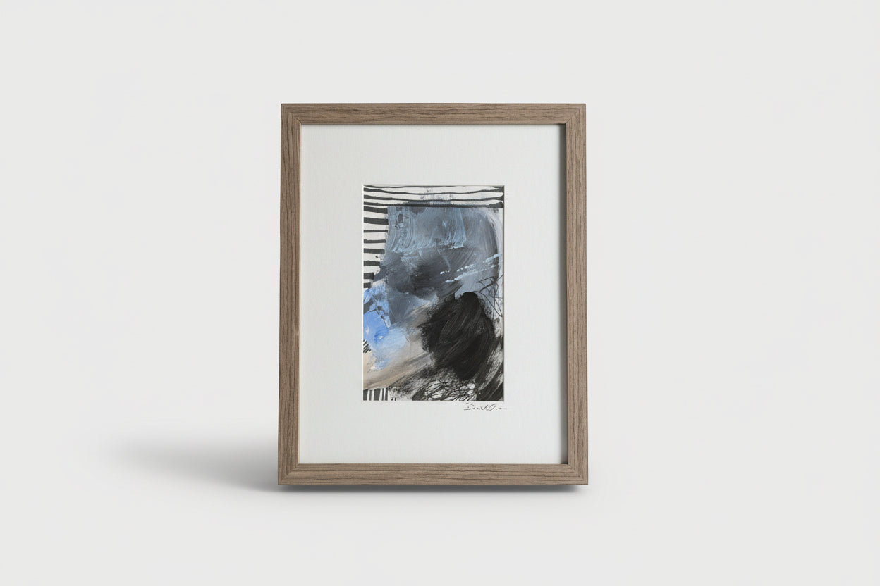 A framed mixed media artwork on a wooden surface, with abstract blue and black colors, placed next to a black vase with a flower.