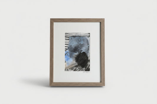 A framed mixed media artwork on a wooden surface, with abstract blue and black colors, placed next to a black vase with a flower.
