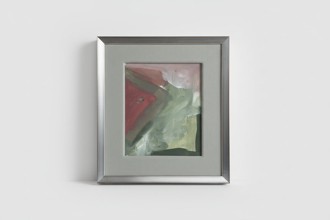 A framed abstract artwork featuring red, green, and beige colors, placed on a wooden surface with a blurred background.