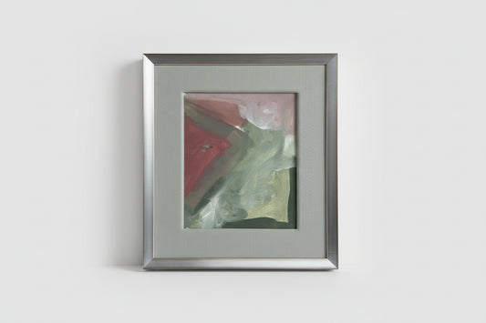 A framed abstract artwork featuring red, green, and beige colors, placed on a wooden surface with a blurred background.