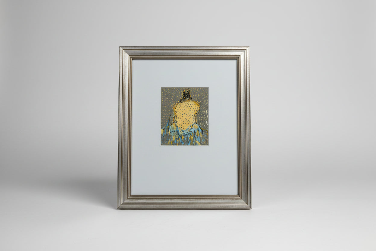A framed mixed media artwork of a woman's figure on paper, displayed on a wooden surface with a decorative item in the background.
