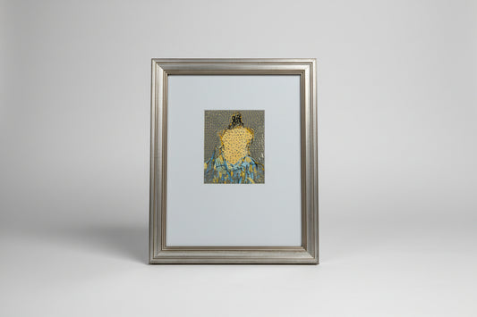 A framed mixed media artwork of a woman's figure on paper, displayed on a wooden surface with a decorative item in the background.