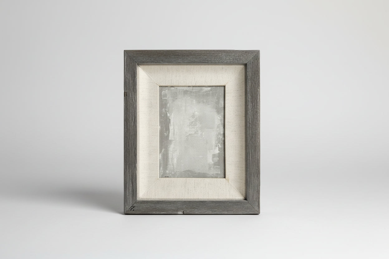 An acrylic on linen panel artwork with a gray wood frame, displayed on a concrete surface with green plants in the background.