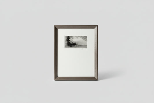 A framed artwork featuring a monochrome scene with a boat on water, placed on a wooden surface with a plant in the background.