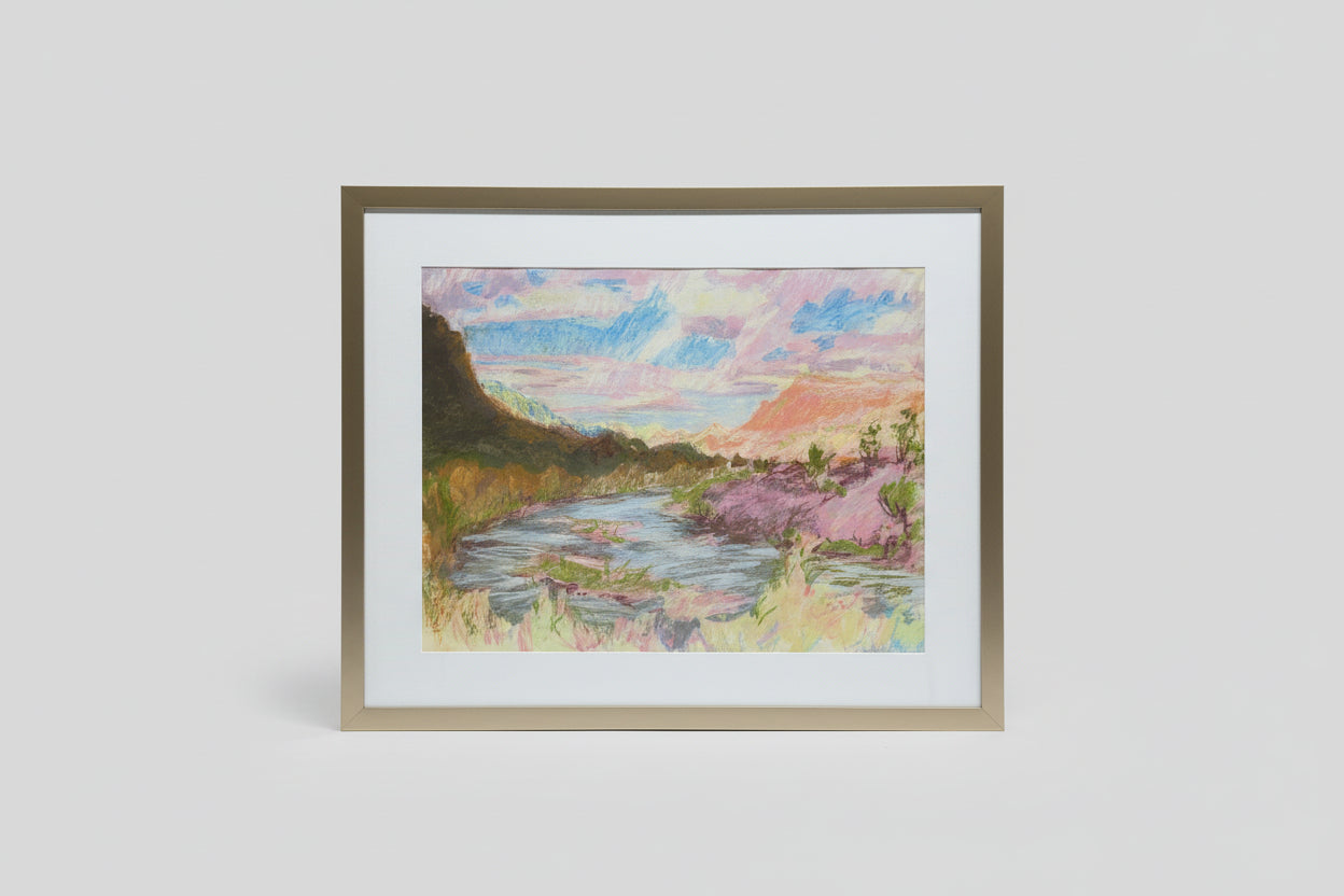 A framed mixed media artwork depicting a peaceful landscape with a horizon, displayed on a wooden surface with green plants in the background.