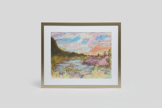 A framed mixed media artwork depicting a peaceful landscape with a horizon, displayed on a wooden surface with green plants in the background.