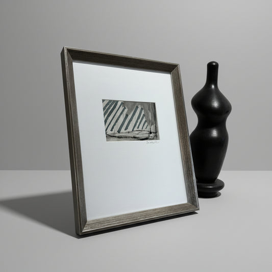 A framed artwork featuring geometric patterns in blue and white, placed on a wooden surface with a vase and sculpture in the background.