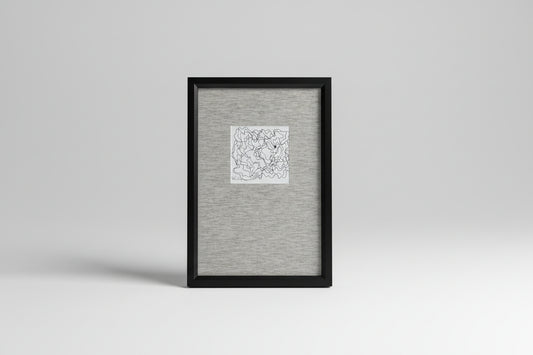 A framed artwork featuring a scribble face drawing, placed on a wooden surface with a plant in the background.