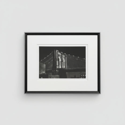 A framed and matted black and white photograph of the Brooklyn Bridge, displayed on a wooden surface.
