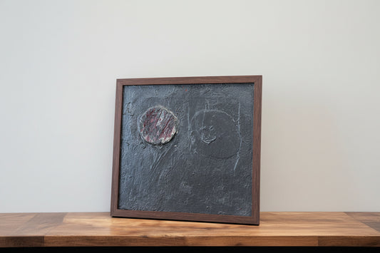 An artwork with heavy oil on canvas, featuring a dark and textured surface, framed in a wooden frame, placed on a wooden shelf next to a black sculpture.
