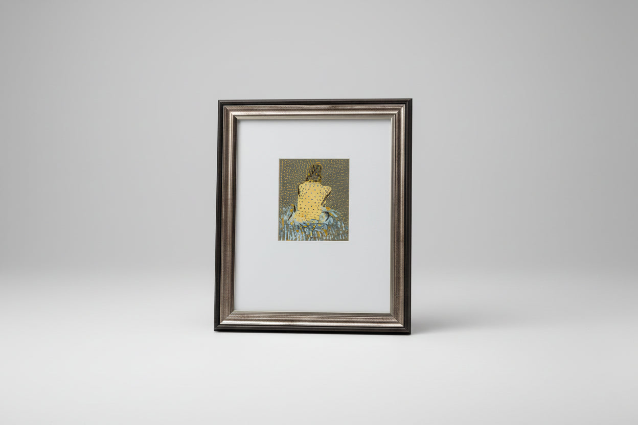 A framed mixed media artwork of a woman's figure, displayed on a wooden table with a sculpture in the background.