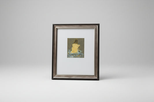 A framed mixed media artwork of a woman's figure, displayed on a wooden table with a sculpture in the background.
