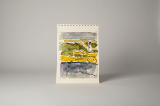 A framed mixed media artwork featuring a lighthouse on paper, displayed on a table with a blurred background of a window and plants.