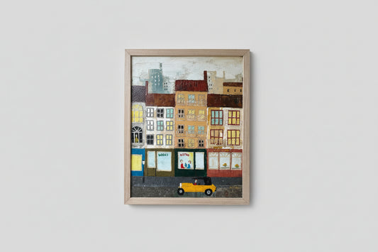 A framed artwork depicting a colorful town scene with buildings and a car, displayed in a well-lit room with a custom wood frame.