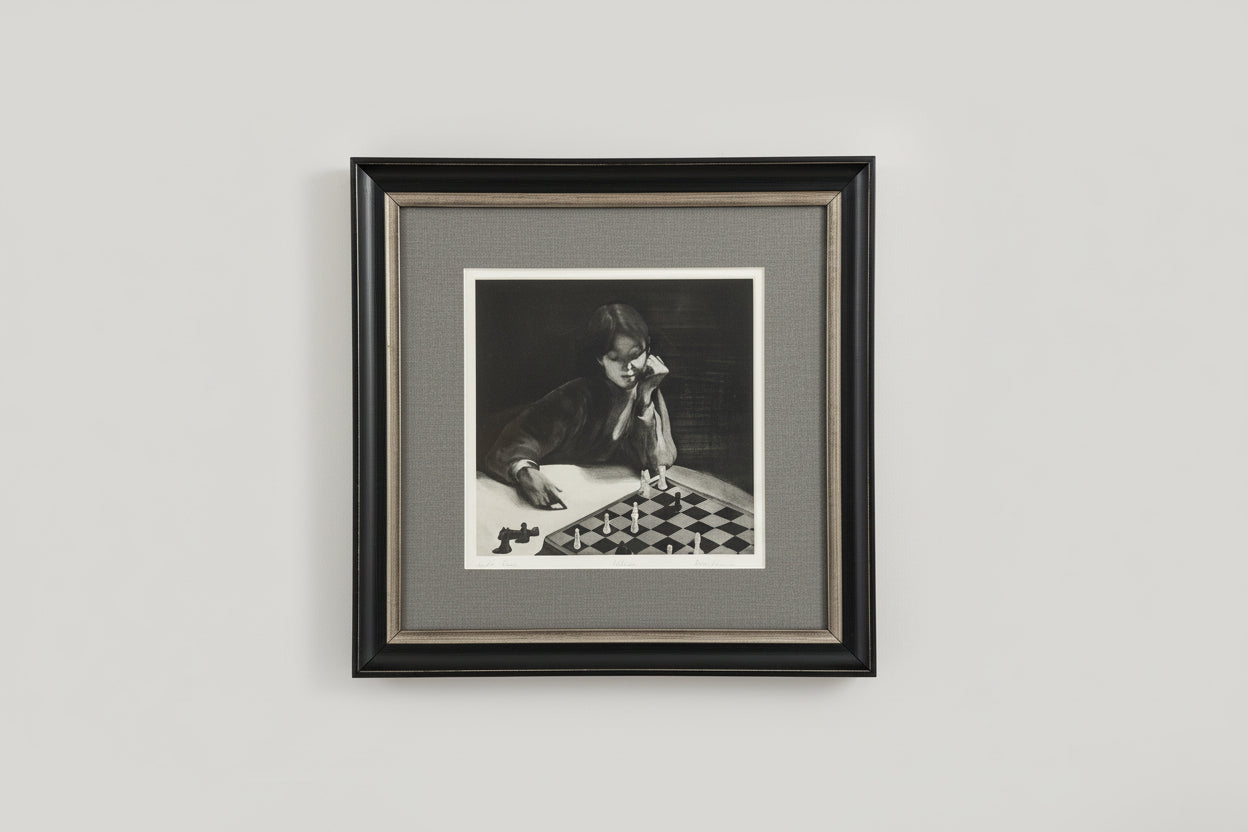 A framed and matted black and white etching of a person sitting at a chess board, displayed on a wall.