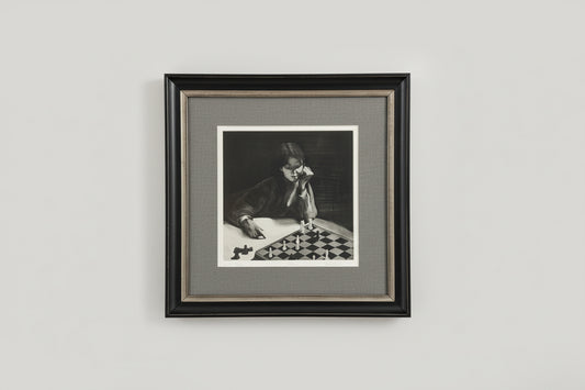 A framed and matted black and white etching of a person sitting at a chess board, displayed on a wall.